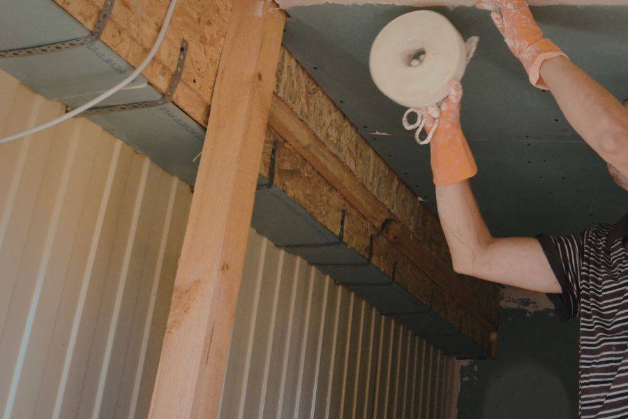 A person wearing orange gloves applying sealing tape to roof insulation panels between wooden beams, as part of insulating your home yourself.