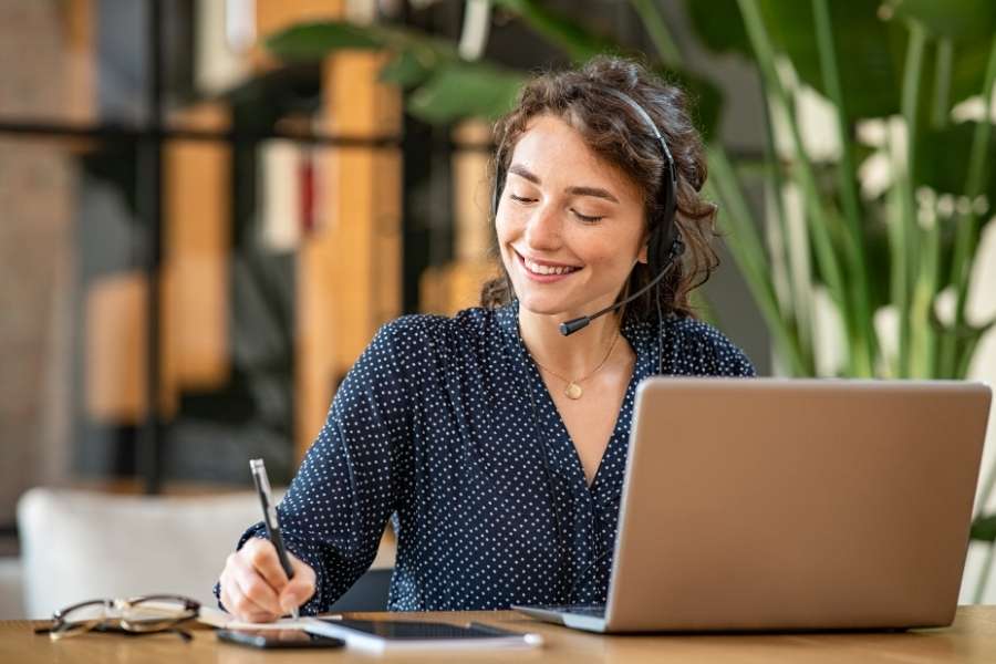 A smiling woman wearing a headset with a microphone writes in a notebook while looking at her laptop on a wooden desk.