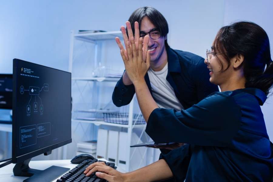 Two smiling colleagues giving a high five in front of a computer screen showing digital configurations.