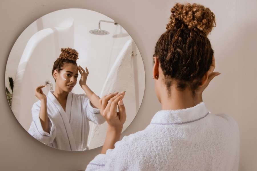 A woman in a white bathrobe looking into a round mirror as part of her evening rituals.