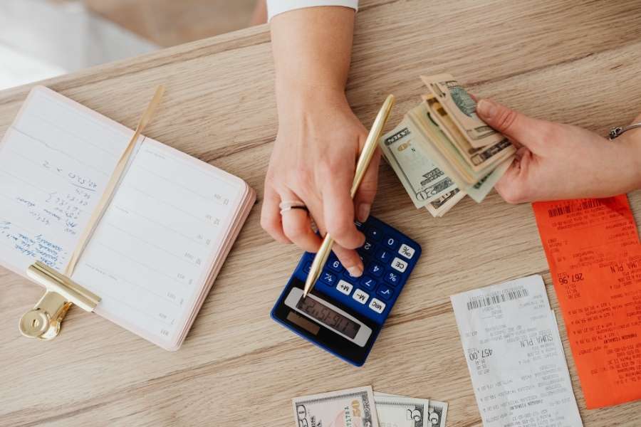 A person using a calculator, handling dollar bills, and reviewing receipts on a wooden table.